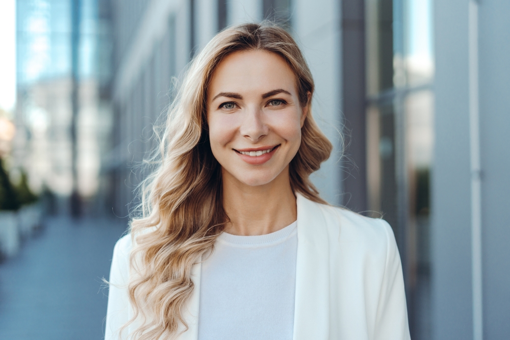 A smiling woman in a white blazer poses in an urban setting, highlighting confidence and clarity enhanced by her fashionable eyeglasses – Eyeglasses in Dearborn.