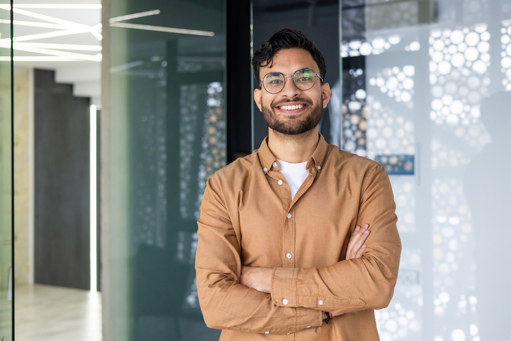 A cheerful man wearing glasses stands confidently with folded arms in a bright, contemporary office setting, reflecting professionalism and modern eyecare values – Eye Doctor in Detroit.