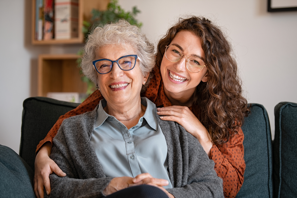 A joyful senior woman and her adult daughter, both wearing stylish eyeglasses, pose together at home, showcasing the importance of lifelong vision care – Eye Doctor in Detroit.