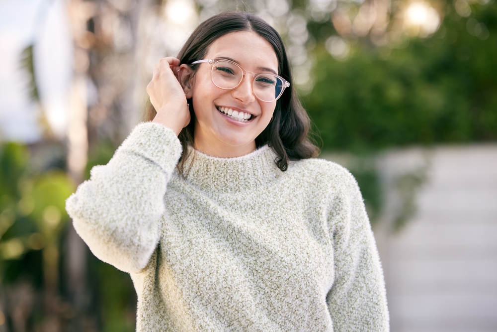 A smiling young woman with clear-framed glasses enjoys a bright outdoor setting, representing confidence and clarity in vision – Eye Doctor in Detroit.